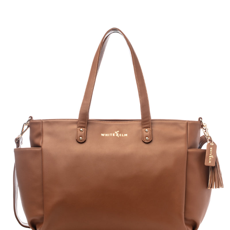 A caramel-colored Aquila tote bag made from vegan leather, featuring two handles, a tassel detail, and side pockets.