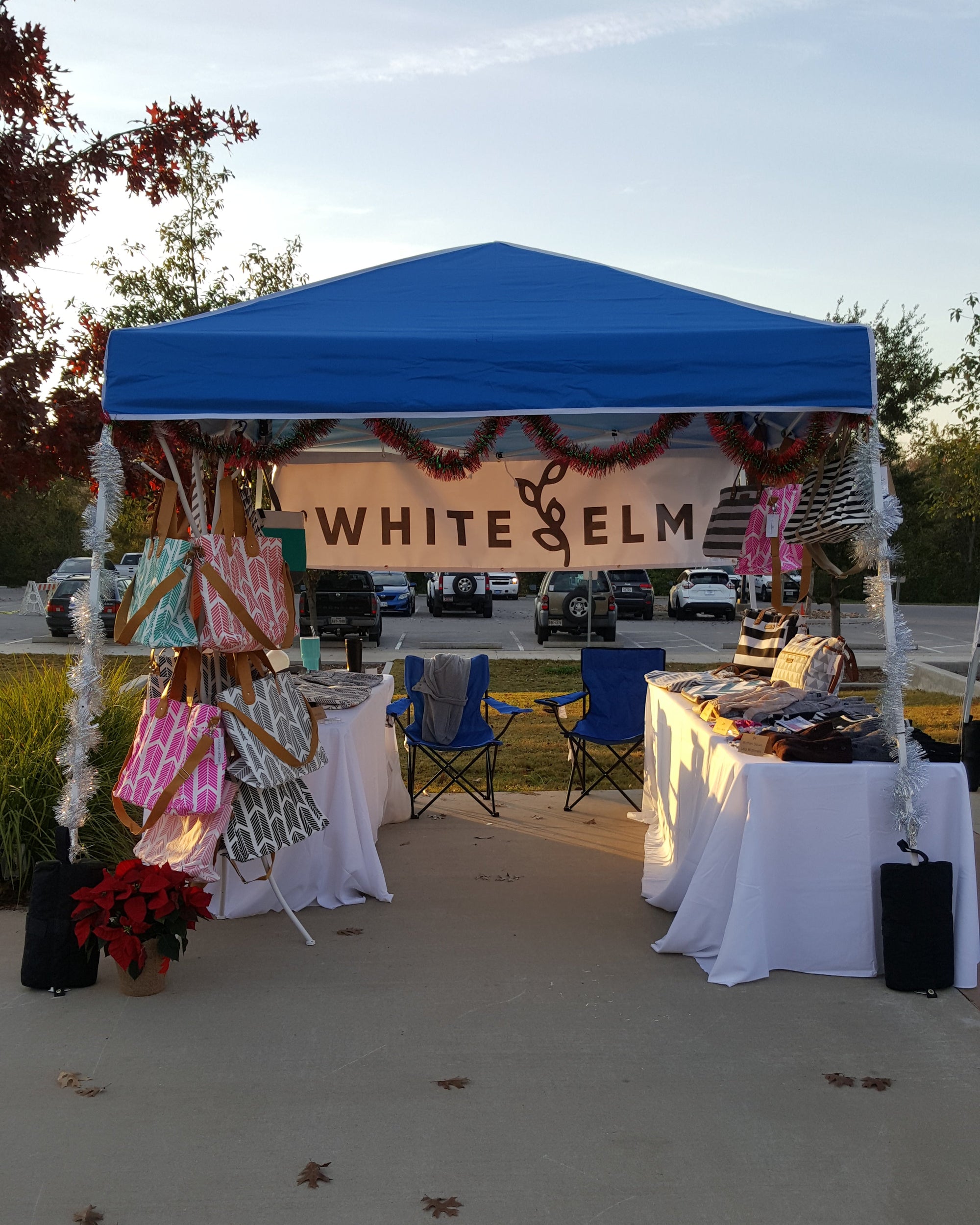 A white outdoor booth under a blue canopy tent with the words 'WHITE ELM' on the front, set up on a concrete surface with tables, chairs, and hanging decorations.