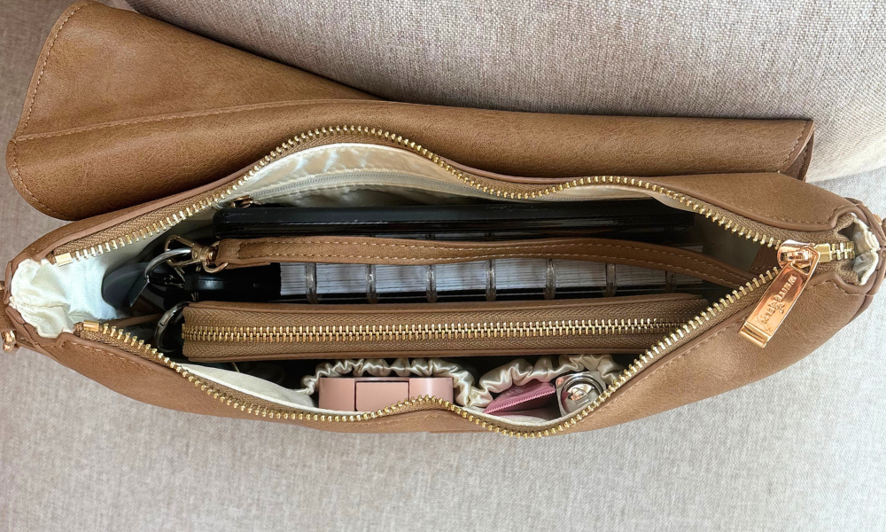 A tan faux-leather cosmetic pouch shown open, revealing its gold-toned zipper, cream interior lining, and packed contents including a black case, clear rectangular items, and pink accessories, set against a neutral textured fabric background.