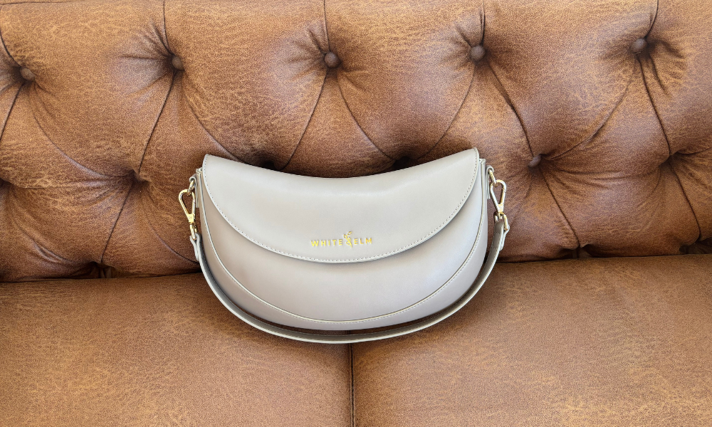 A light beige leather crescent-shaped shoulder bag with gold hardware and a small gold logo on the front, resting on a brown tufted leather sofa.