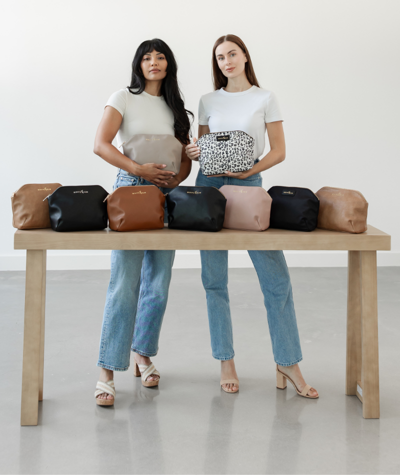 Two women standing behind a wooden table displaying a collection of small zip pouches in multiple colors, including beige, black, brown, pink, and leopard print.