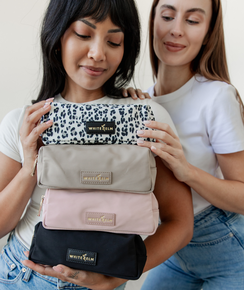 Two women holding a stack of four White Elm Penny Pencil pouches in different colors: black leopard print, taupe, dusty pink, and black.