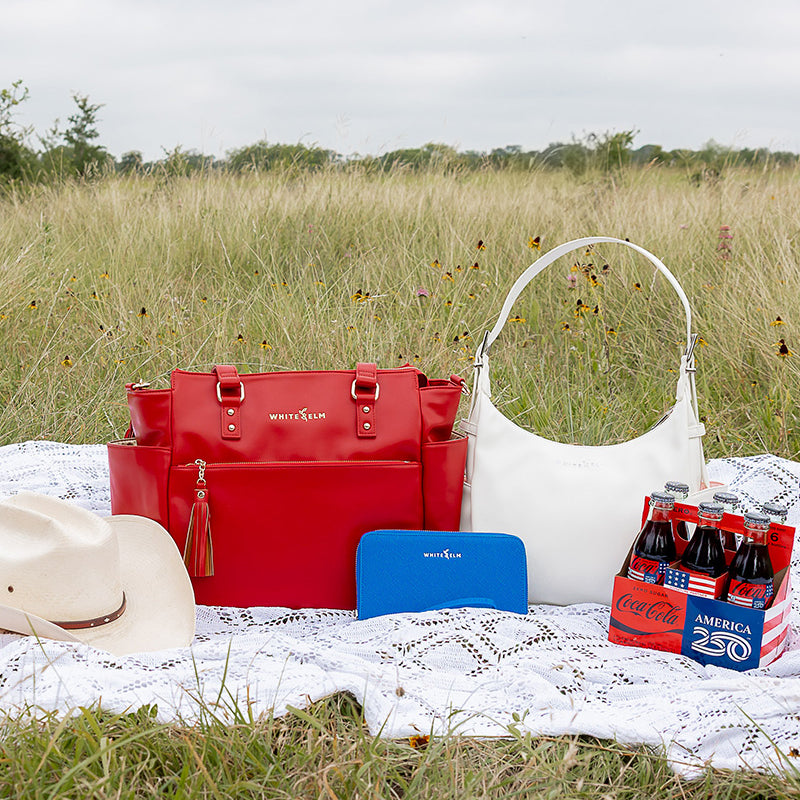 Red white and blue handbags on a picnic blanket with Coca-Cola bottles in a field.