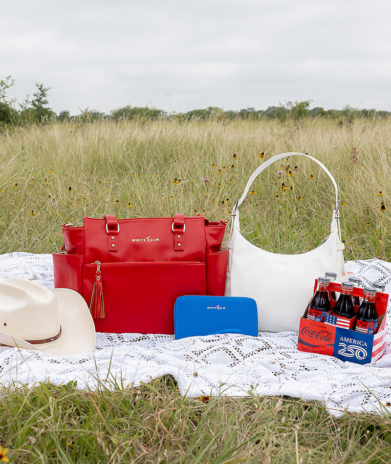 Red white and blue handbags on a picnic blanket with Coca-Cola bottles in a field.