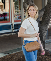A woman wearing a white short-sleeve mock neck top and blue jeans carries a tan leather crossbody bag with a matching strap, standing outdoors next to a tree.
