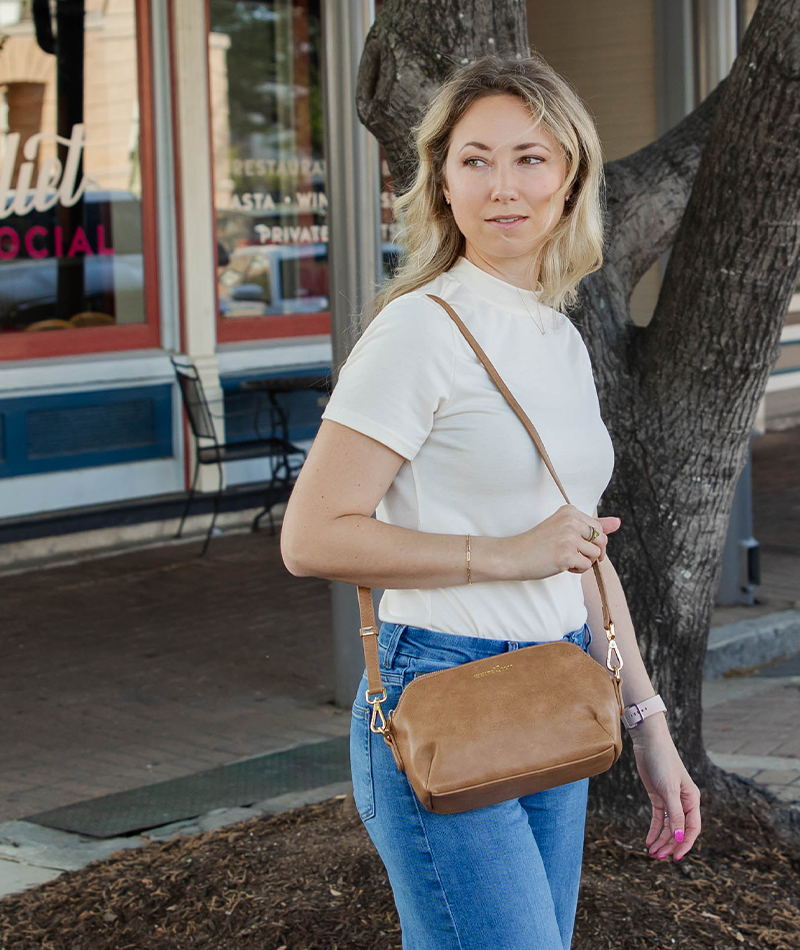 A woman wearing a white short-sleeve mock neck top and blue jeans carries a tan leather crossbody bag with a matching strap, standing outdoors next to a tree.