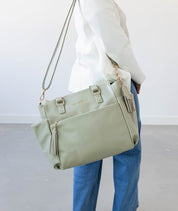 Person holding a light sage green tote bag with gold accents on a plain background