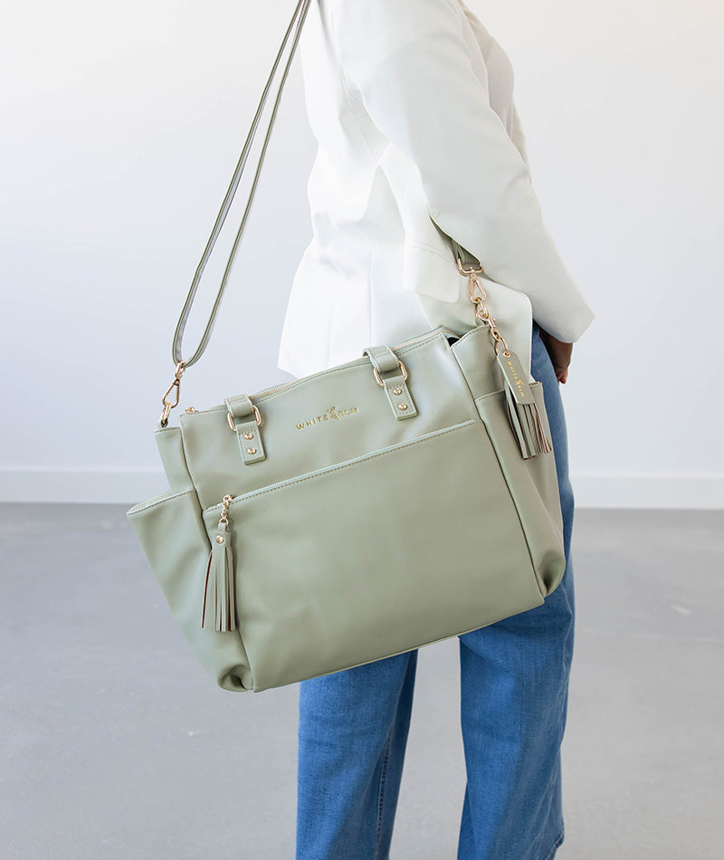 Person holding a light sage green tote bag with gold accents on a plain background