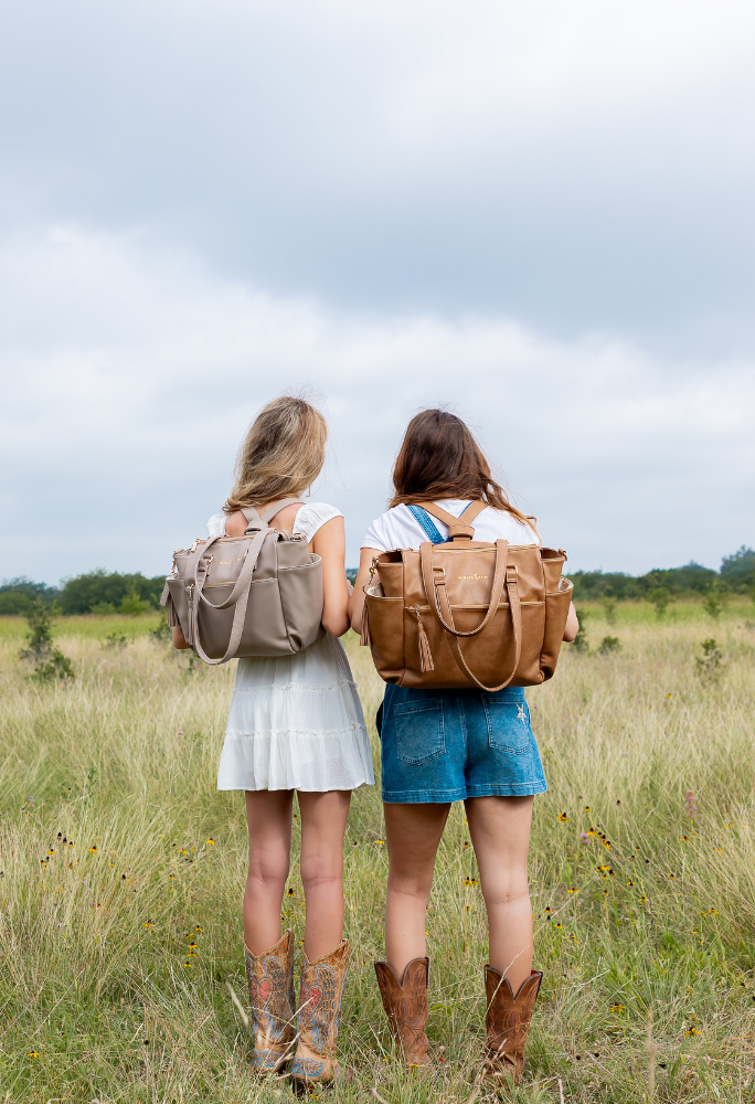 Two women wearing White Elm Gemini backpacks in taupe and caramel, standing in a grassy field
