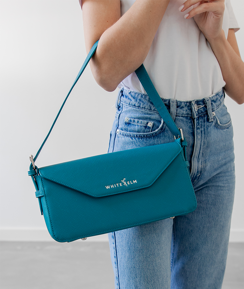 Woman carrying the teal Noemi shoulder bag on her forearm in a white top