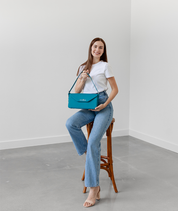 Smiling woman seated on a stool holding the teal Noemi east west bag by its strap