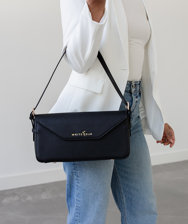 Woman carrying the black Noemi east west bag on her forearm in a white blazer