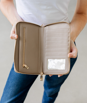 An open taupe zip-around wallet with gold-tone hardware, held open by a person wearing a white t-shirt and blue jeans. The wallet features multiple card slots, a vertical zip pocket, and a clear ID window.